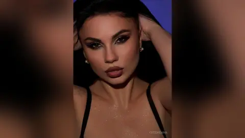 CutemEmma poses with her hands in her dark hair, wearing a black camisole or bra top against a blue background.