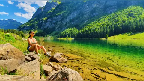 Young woman in casual summer attire sitting on rocks by a serene mountain lake under a bright blue sky.