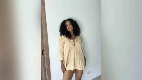 Curvy woman with curly hair wearing a beige oversized button-down shirt in a bright, minimalist room.