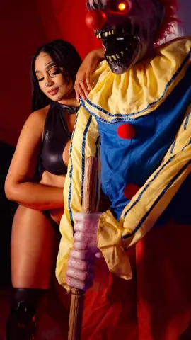 Woman in black bodysuit posing with a creepy clown character in a colorful costume against a red background.