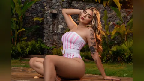 Blonde woman in a pink striped swimsuit relaxing on a wooden deck in a lush tropical garden setting.