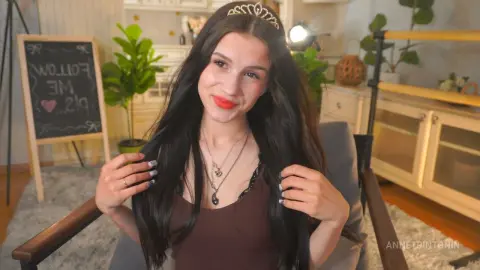 Smiling young woman with long dark hair and a tiara wearing a brown top in a bright, cozy room.