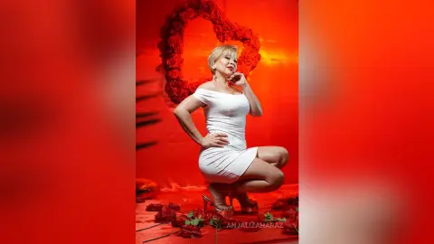 AnjaliZaharaz poses in a tight white dress and red high heels against a red heart-shaped rose backdrop.