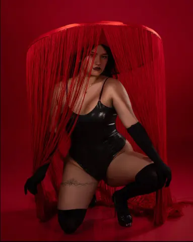 Glamorous woman in a black bodysuit and gloves posing under red fringe curtains against a red background.