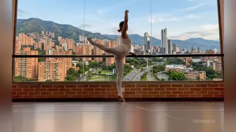 TefyRusso poses in a white leotard, performing a ballet stretch with one leg raised high against a city and mountain backdrop.