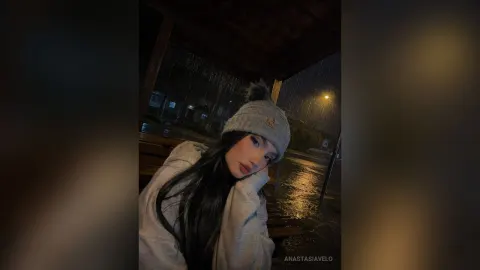 AnastasiaVelo — AnastasiaVelo wears a grey beanie and a cozy white sweater while sitting on a wooden bench in the rain at night.