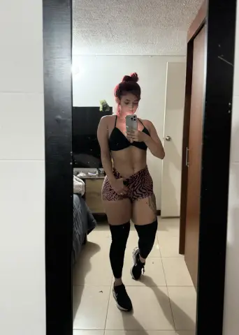 RebbecaWells takes a mirror selfie wearing a black bikini top and pink patterned shorts while posing in her bedroom.