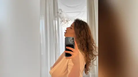 Young woman in a white blouse taking a mirror selfie in a bright, airy room with soft sunlight.
