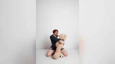 AryaSin, a redheaded woman, kneels on a white floor while hugging a large tan teddy bear, wearing a dark long-sleeved shirt and dark shorts.