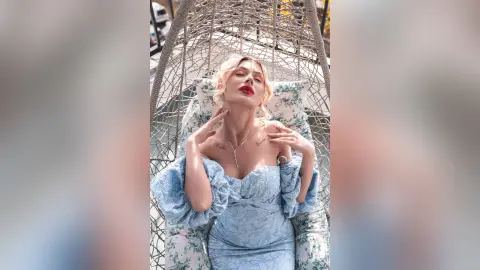 DaisyLin leans back in a hanging wicker chair wearing a light blue off-the-shoulder dress with red lipstick.
