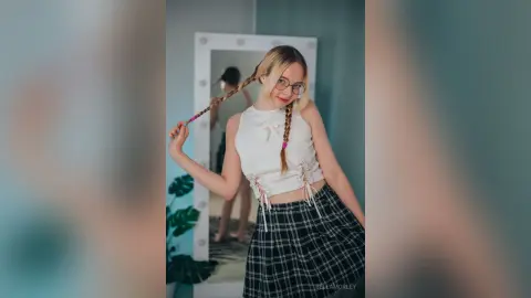Young woman with long braids in a white crop top and plaid skirt posing in front of a vanity mirror.