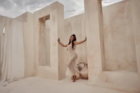 Elegant woman in a white flowing dress posing against minimalist beige stone walls in a dreamy desert setting.