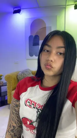 Young woman with long dark hair wearing a white and red graphic t-shirt in a cozy living room setting.