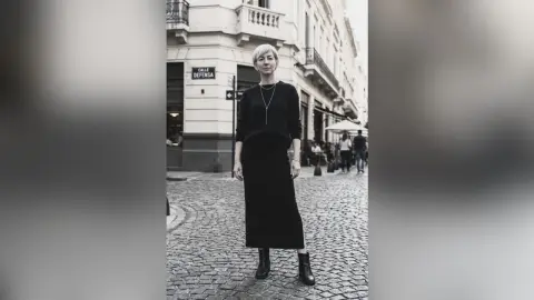 BellaMontclaire stands on a cobblestone street wearing a long black dress and boots, looking at the camera.