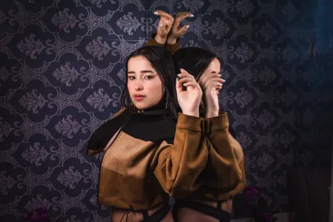 Young woman in a brown suede jacket posing against dark damask wallpaper in a moody, cinematic setting.