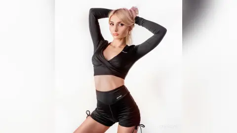 MonaLys poses with her hands behind her head, wearing a black long-sleeve wrap top and tight black athletic shorts against a white background.