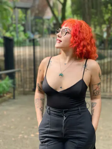 Woman with red hair and tattoos wearing a black bodysuit and necklace in a lush outdoor garden setting.
