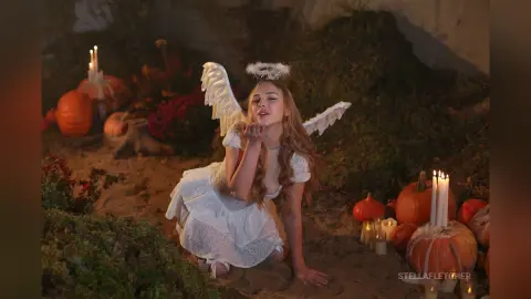 StellaFletcher wears white lace angel wings and a white lace dress while kneeling on sand surrounded by lit candles and pumpkins.