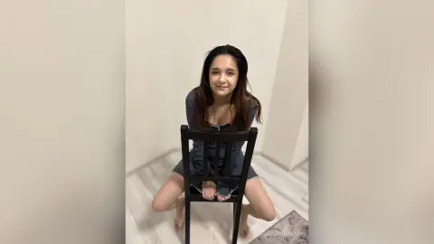 Smiling young woman in a casual dark blue dress sitting on a black chair in a bright minimalist room.
