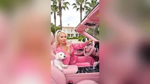 Blonde woman in pink loungewear relaxing in a luxury pink convertible car with a small white puppy.