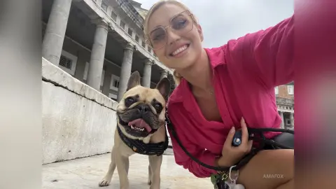 AmaFox — Smiling blonde woman in a pink shirt taking a selfie with her cute French Bulldog in front of a classical building.