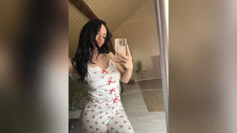 Smiling woman in white floral pajama set taking a mirror selfie in a bright, cozy bedroom.