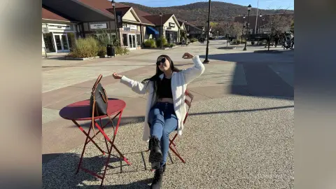 Cheerful woman in a white cardigan and jeans standing by a red table in a sunny outdoor shopping plaza.