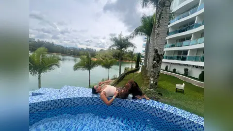 Relaxed woman in a beige top lounging by a blue tiled pool at a tropical luxury resort under a cloudy sky.
