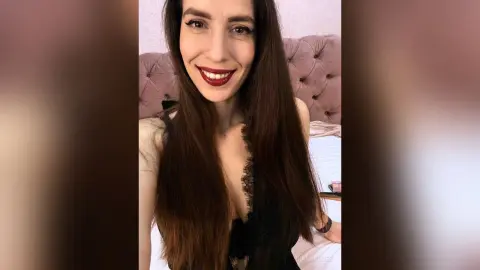 Smiling brunette woman in a black lace dress posing in a cozy bedroom setting.