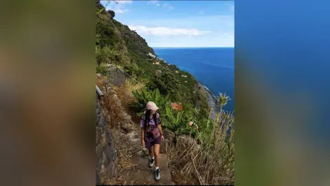 Hiker in a pink hat and backpack walking along a scenic coastal mountain trail overlooking the blue ocean.