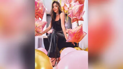 JessieLiu poses in a black tank top and black pants, surrounded by pink and gold balloons.