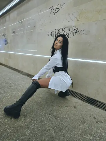 Stylish woman in a white shirt and black boots posing against a graffiti wall in an urban subway setting.