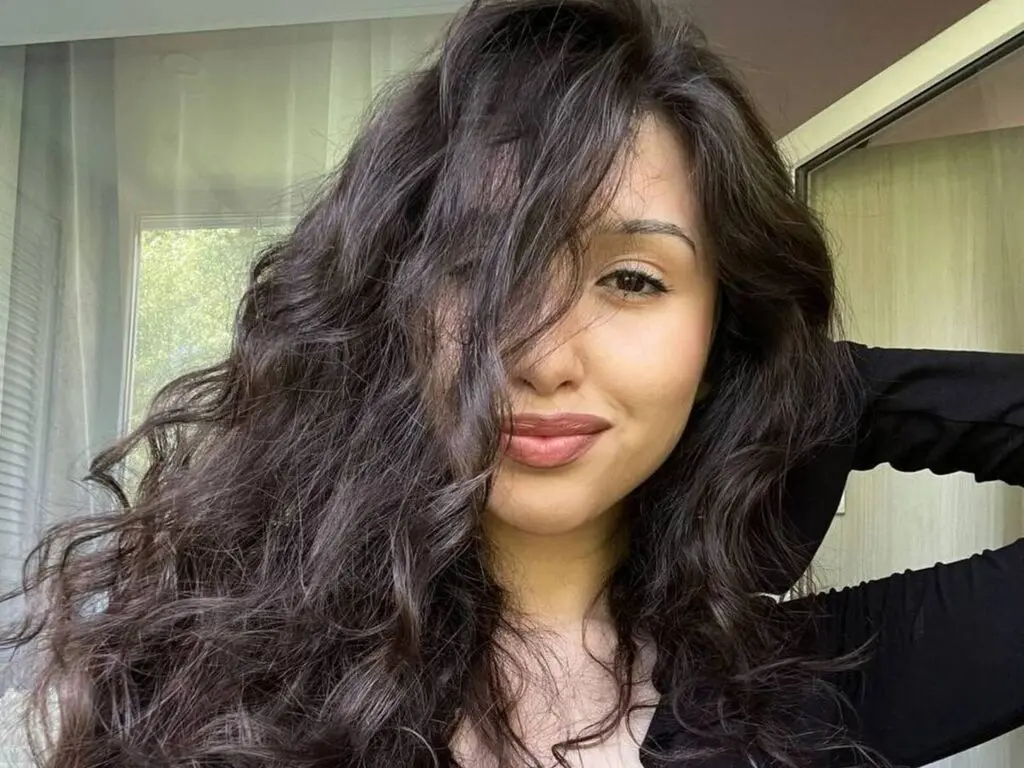 Woman with long wavy brunette hair wearing a black top in a bright, natural indoor setting.