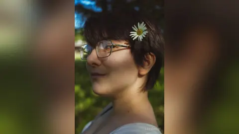 Smiling young woman with short hair and a daisy tucked behind her ear in a sunny outdoor setting.