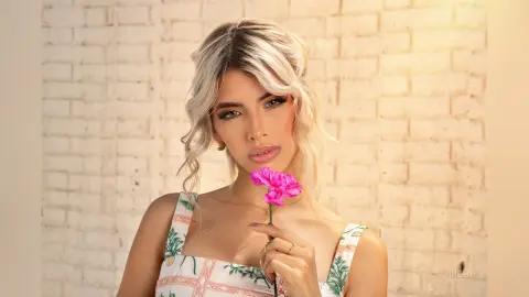 Blonde woman in a floral white dress holding a pink flower against a white brick wall in soft natural light.