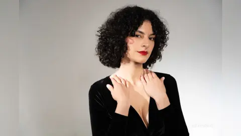 RoxyGraff poses with her hands on her neck, wearing a black velvet top with a deep v-neckline against a plain grey background.