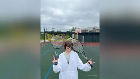 Young woman in a white hoodie holding tennis rackets on an outdoor court under a cloudy sky.