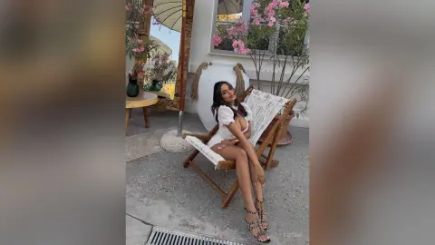 Smiling woman in a white summer dress sitting on a lounge chair in a sunny Mediterranean garden setting.