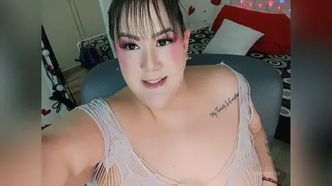 Woman with pink makeup and white lace top smiling in a cozy bedroom setting.
