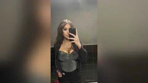 Young woman in a sparkly grey corset top and black jacket taking a moody mirror selfie in a dark bathroom.
