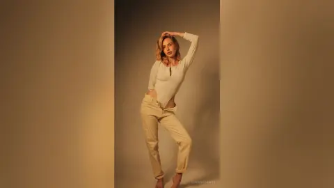 Blonde woman in a white bodysuit and beige trousers posing against a neutral studio background with warm lighting.