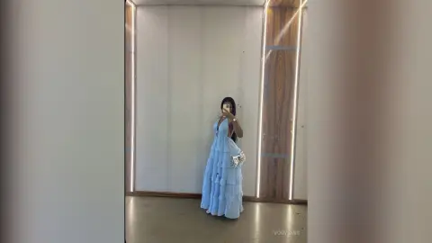 Woman in a flowing light blue maxi dress taking a mirror selfie in a modern minimalist room.