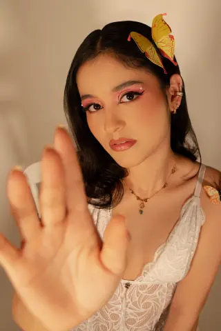 Young woman in white lace camisole with butterfly hair accessory and soft pink makeup in warm lighting.