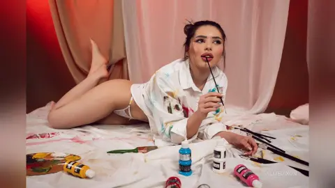 Young woman in a white shirt lying on a messy art canvas surrounded by paint supplies in a soft studio setting.