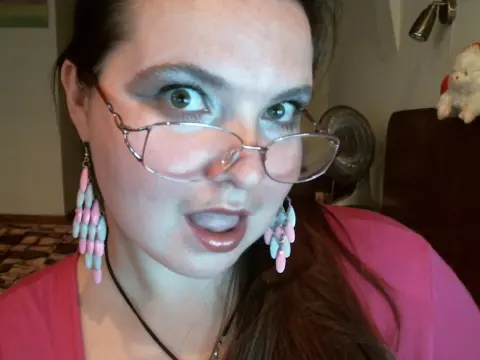 Woman with glasses and pink tassel earrings wearing a pink shirt in a cozy indoor setting.