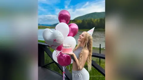 StellaFletcher smiles while holding a pink frosted cake next to a large bunch of pink and white balloons outdoors.