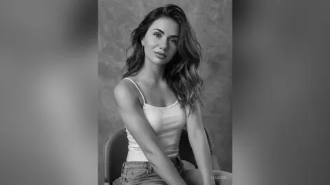 Portrait of a smiling young woman in a white tank top posing against a textured wall in black and white.