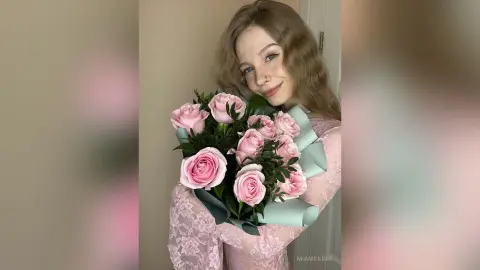 Smiling young woman in a pink lace dress holding a large bouquet of fresh pink roses in a bright indoor setting.