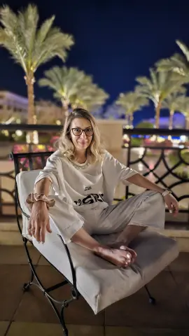 Relaxed woman in white loungewear sitting on a patio chair under palm trees at night.