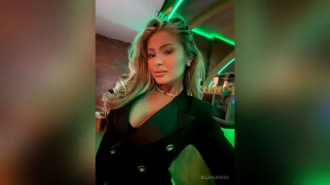Glamorous blonde woman in a chic black blazer posing in a moody, neon-lit nightclub setting.